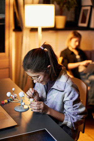 Vertical shot of girl sitting at desk working on solar system model, her mother relaxing on sofa...