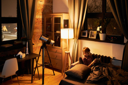 Long shot of girl wearing eyeglasses relaxing in bed in evening reading book about solar system before going to sleepの写真素材