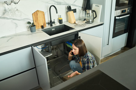 Caucasian young adult woman crouching under kitchen sink talking on smartphone appearing concerned while inspecting plumbing pipes in modern kitchen interiorの写真素材