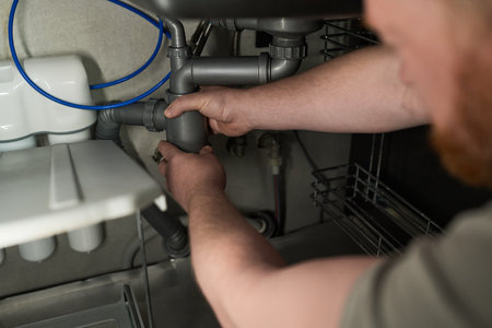 Caucasian middle aged man repairing kitchen sink plumbing under counter, using both hands to adjust...