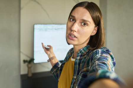 Portrait of young adult Caucasian woman looking into camera while holding remote control and...