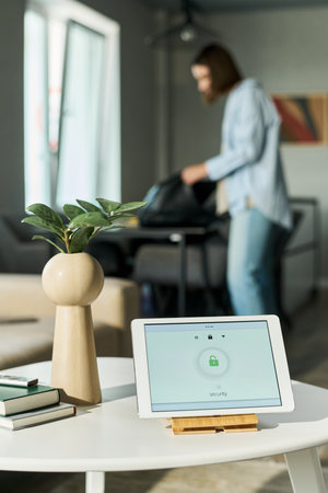 Caucasian young adult woman standing in modern living room packing bag in background, digital tablet with security lock screen displayed on table in foreground, plant and books nearbyの写真素材