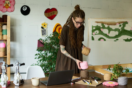 Young adult Caucasian woman with tattoos arranging yarn balls on worktable in carpet workshop,...