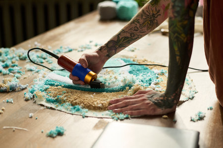 Tattooed young adult Caucasian man using electric tufting gun creating handmade carpet on wooden...