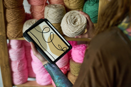Tattooed young adult Caucasian woman holding ball of yarn in one hand and digital tablet displaying...