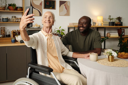 Senior Caucasian woman in wheelchair holding smartphone taking selfie with young Black man sitting...