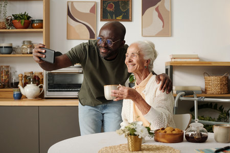 Young Black man taking selfie with senior Caucasian woman holding cup, both smiling at kitchen table...