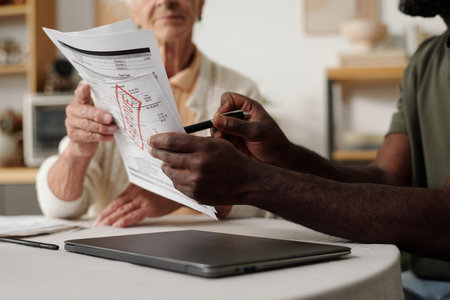 Senior Caucasian woman holding document while Black man pointing at printed chart, both sitting at...