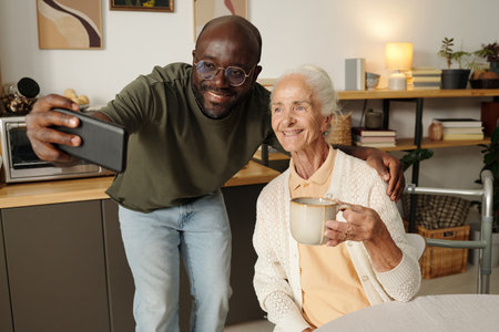 Young Black man smiling while taking selfie with senior Caucasian woman holding mug, both standing...