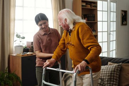 Young adult Latin woman assisting senior Caucasian man using walker in living room, both smiling and...