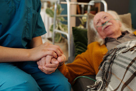 Senior Caucasian man lying in bed with oxygen tube holding hands with young adult woman caregiver,...