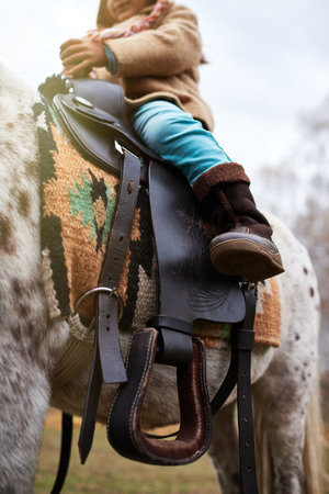 Child sitting on horseback, holding reins, wearing winter clothing, visible lower body and hands,...