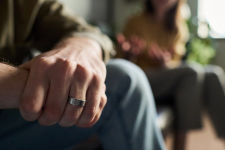 Caucasian young adult man wearing wedding ring sitting with hand in focus, while blurred young adult...