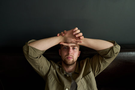 Portrait of young adult Caucasian man reclining on dark sofa, gazing directly into camera with...