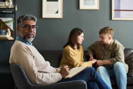 Portrait of middle aged Indian man with beard looking into camera while holding notebook, sitting in...