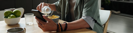 Young adult Caucasian man sitting at table using smartphone while pouring milk into bowl of cereal,...