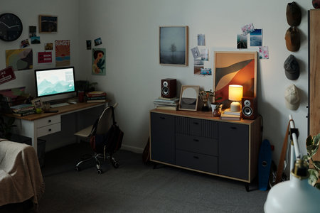 Teenager sitting at desk using computer in bedroom decorated with posters and photos, digital device screen glowing, suggesting gadget addiction and prolonged screen timeの写真素材