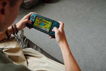 Teenager boy with light skin holding handheld gaming console playing video game, sitting indoors,...