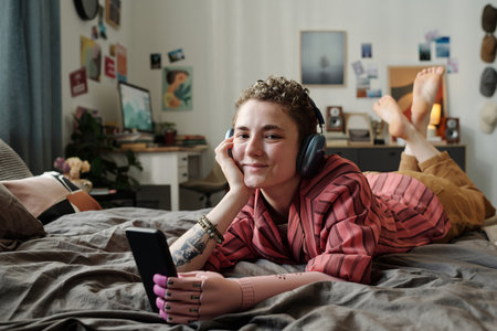 Portrait of young adult Caucasian woman with prosthetic arm lying on bed wearing headphones smiling...