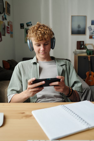 Teenage Caucasian boy wearing headphones sitting indoors using smartphone with focused expression,...
