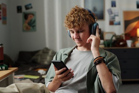Caucasian teenage boy sitting indoors using smartphone while wearing headphones, focusing on screen...