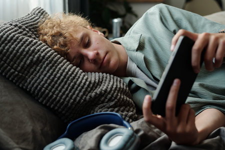 Teenage Caucasian boy lying on bed staring at smartphone with tired expression, holding device close...