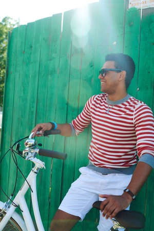 Young adult Hispanic man smiling while sitting on bicycle outdoors, resting one hand on handlebar...