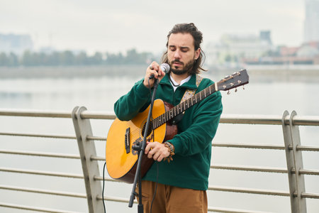 Caucasian young adult man adjusting microphone while holding acoustic guitar outdoors near...