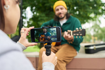Caucasian young adult man playing acoustic guitar outdoors while being recorded on smartphone by...