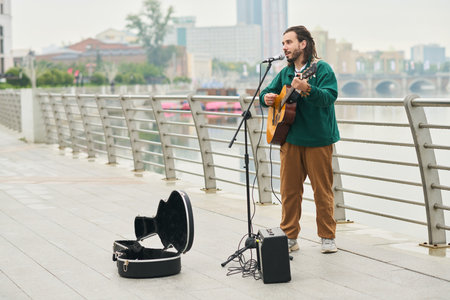 Young adult Caucasian man singing and playing acoustic guitar on city riverside promenade, standing...