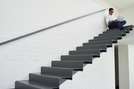 Young adult man sitting on modern staircase working on laptop, focusing on screen, wearing glasses,...