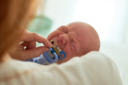 Caucasian infant crying while being comforted by adult woman holding pacifier near mouth, closeup showing newborn facial expression and gentle caregiving interactionの写真素材