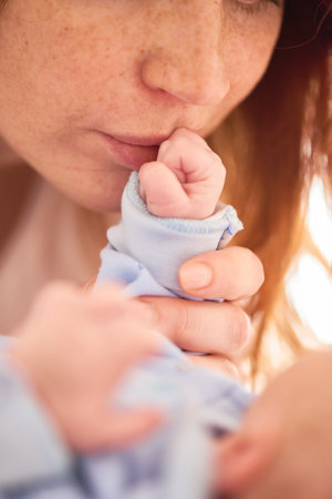 Caucasian young adult woman gently kissing newborn baby hand, showing close up of mother and infant...