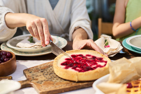 Caucasian young adult man slicing cherry cheesecake with knife while sitting at table outdoors,...
