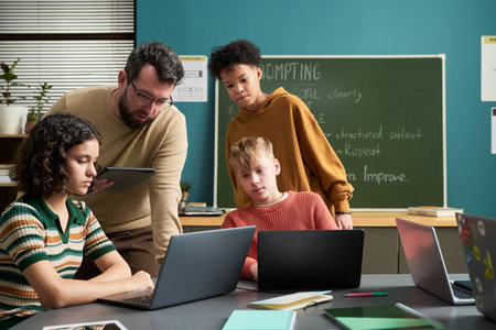 Caucasian man assisting diverse group of teenagers working on laptops in classroom, students...