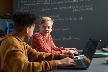 Two teenage boys, one Black and one Caucasian, sitting at desk using laptops, collaborating on coding project in classroom with chalkboard covered in programming code in backgroundの写真素材