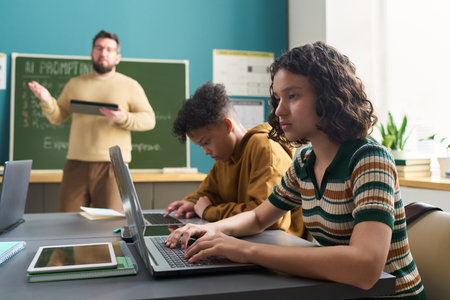 Teenage Caucasian girl and teenage Black boy sitting at desks using laptops in classroom, while male...