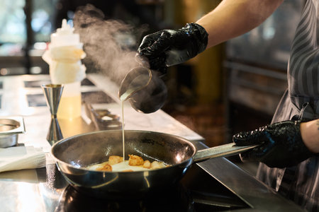 Caucasian young adult man wearing black gloves cooking scallops in frying pan, pouring sauce over...