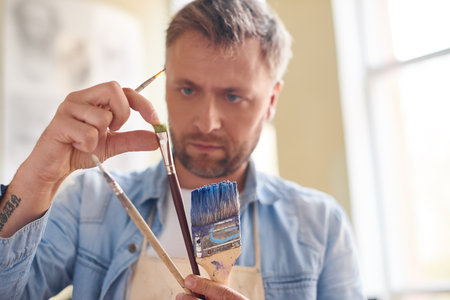 Getting ready for painting. Concentrated fair bearded artist holding paintbrushes of different sizes and checking its qualityの写真素材
