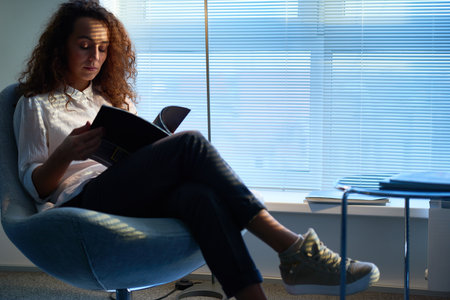 Pretty curly woman wearing white shirt and beige sneakers sitting in waiting room with Venetian blinds on windows, leafing through pages of fashion magazineの写真素材