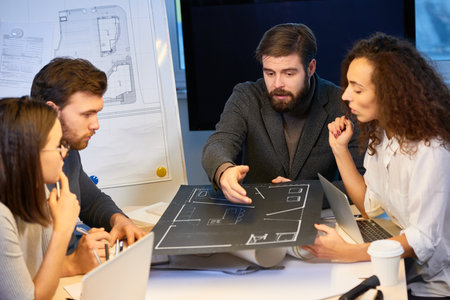Experienced designers sitting at desk in office, holding new house layout made on black wove paper....