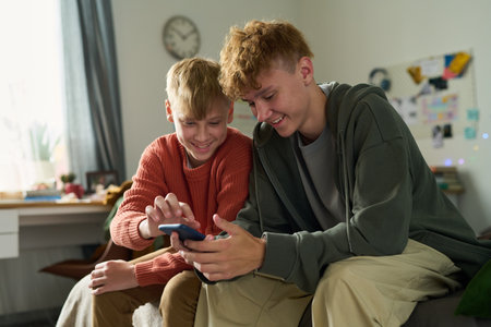 Caucasian teenage boy and Caucasian child boy sitting together on sofa smiling and interacting with...