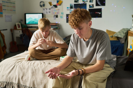 Two Caucasian teenage boys sitting in bedroom, one counting coins in hand while other examining small object, both focused on activity, casual setting with personal items visibleの写真素材