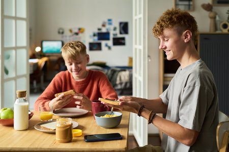 Two Caucasian teenage boys sitting at table eating breakfast together, smiling and holding toast, smartphone and breakfast foods visible on table, casual home settingの写真素材