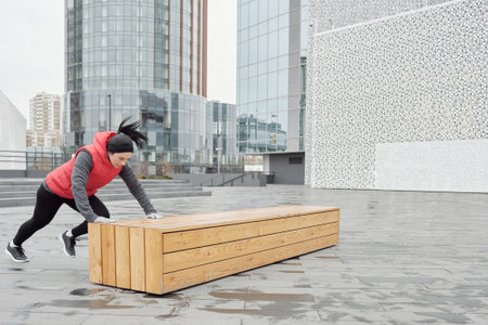 Determined Caucasian woman doing warming-up exercises on wooden bench while having outdoor workout...
