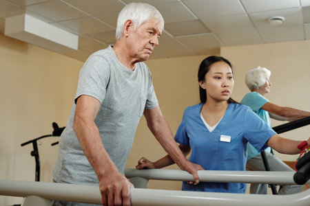 Aged Caucasian man walking on treadmill under control of young female nurse while getting through...