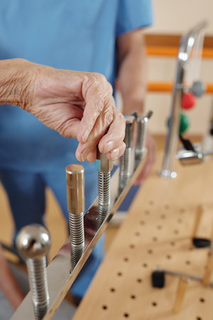 Close-up view of anonymous elderly woman exercising motor skills on training equipment in stroke rehab center under control of physiotherapistの写真素材