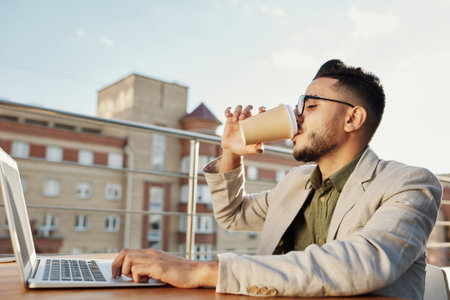 Young attractive Middle Eastern man wearing glasses drinking coffee from paper cup white sitting at table with laptop on rooftop cafe terraceの写真素材