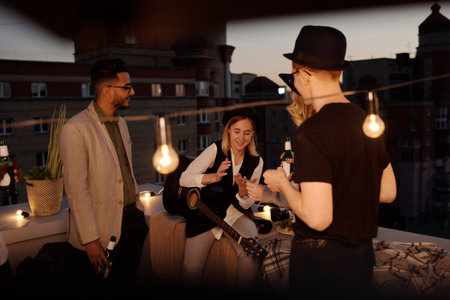Multiethnic group of young stylish people spending summer evening on rooftop, playing the guitar, drinking beer and talkingの写真素材