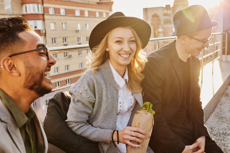 Group of young stylish friends sitting on rooftop terrace with snacks, chatting and enjoying evening, blond pretty woman smiling at cameraの写真素材